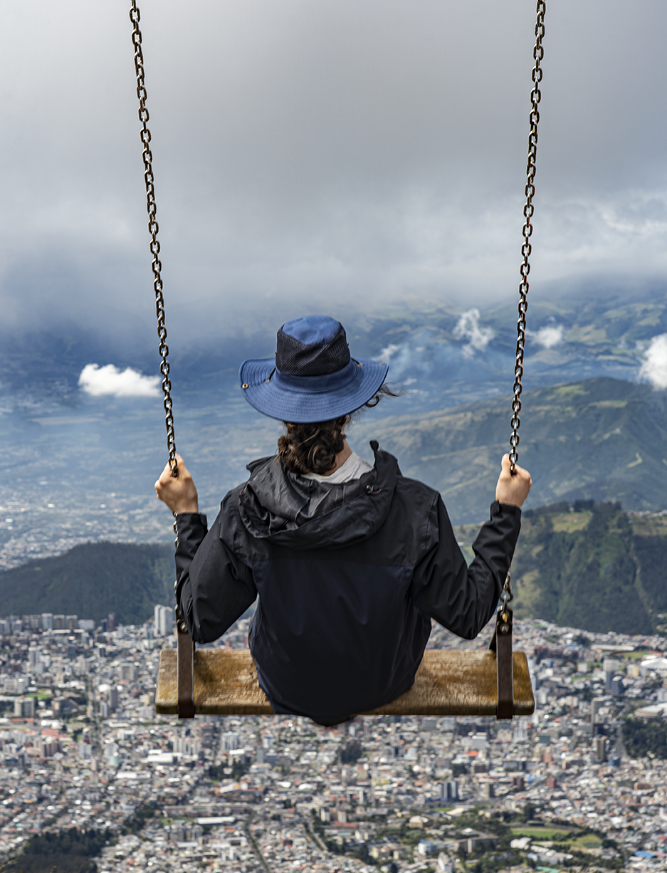 a student sits on a plank swing and swings out on a mountainside, a view of Quito far below them. 