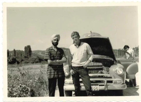 A 1960 photo of Shergill standing with another man in front of a 1960s car on the side of the road with the hood up. 