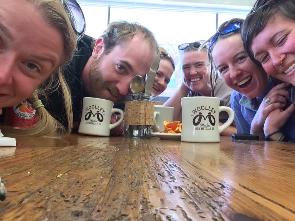 A selfie with six smiling people at a wooden table with coffee cups 