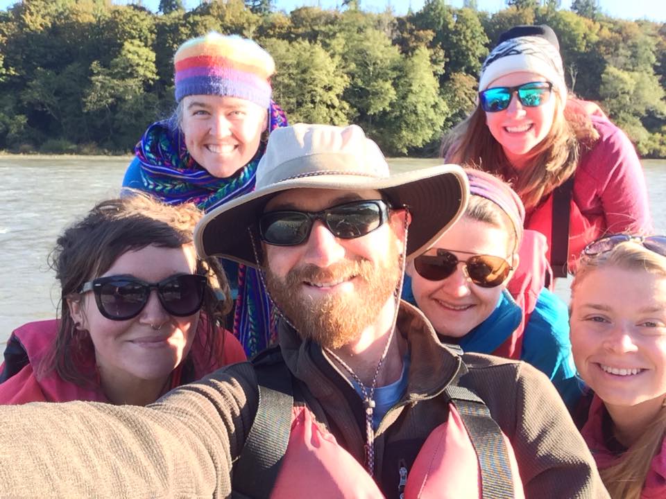 A group selfie of six smiling people by a river on a sunny morning.