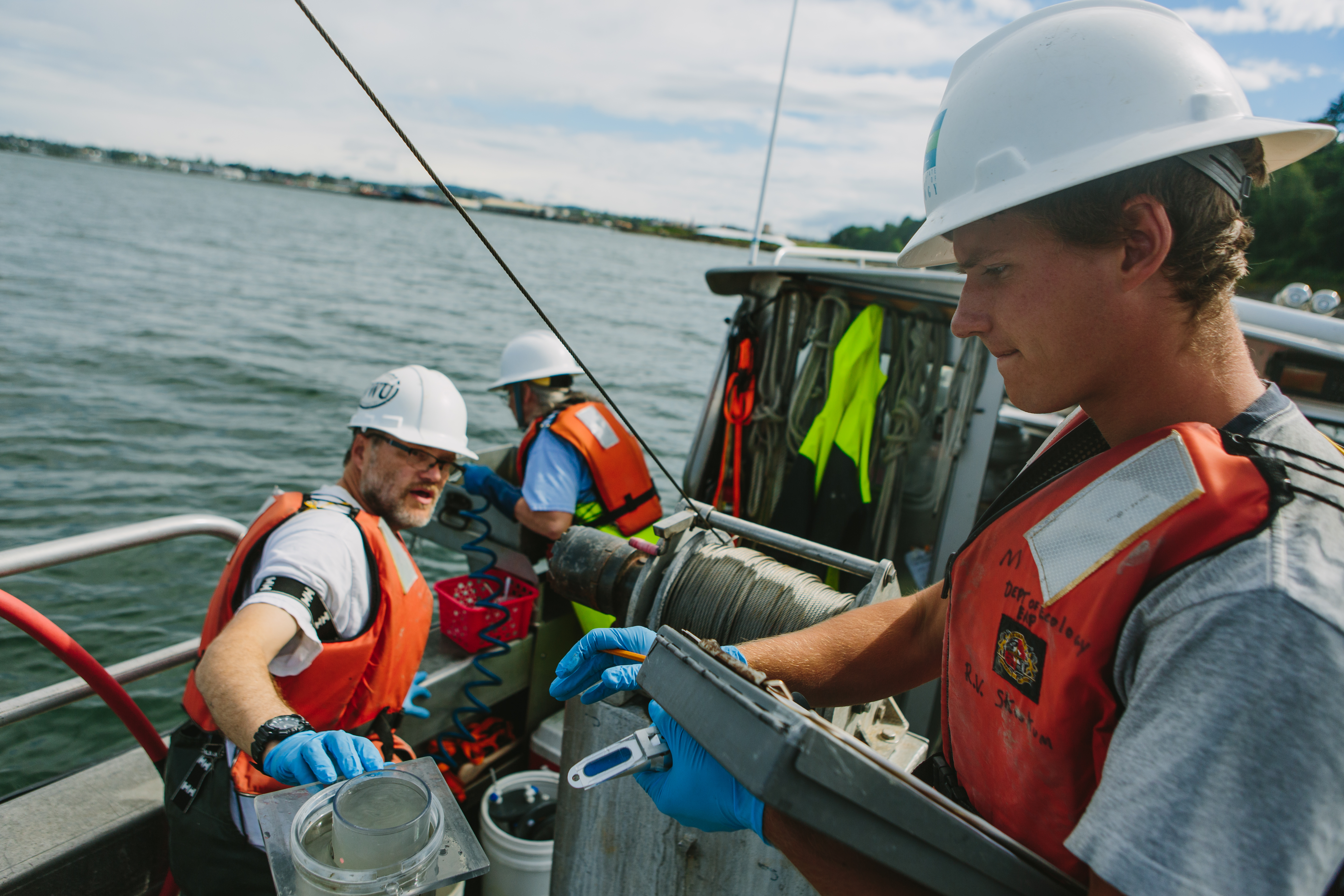a student holds a clipboard while a faculty member hands him a sample jar on a boat in Bellingham Bay
