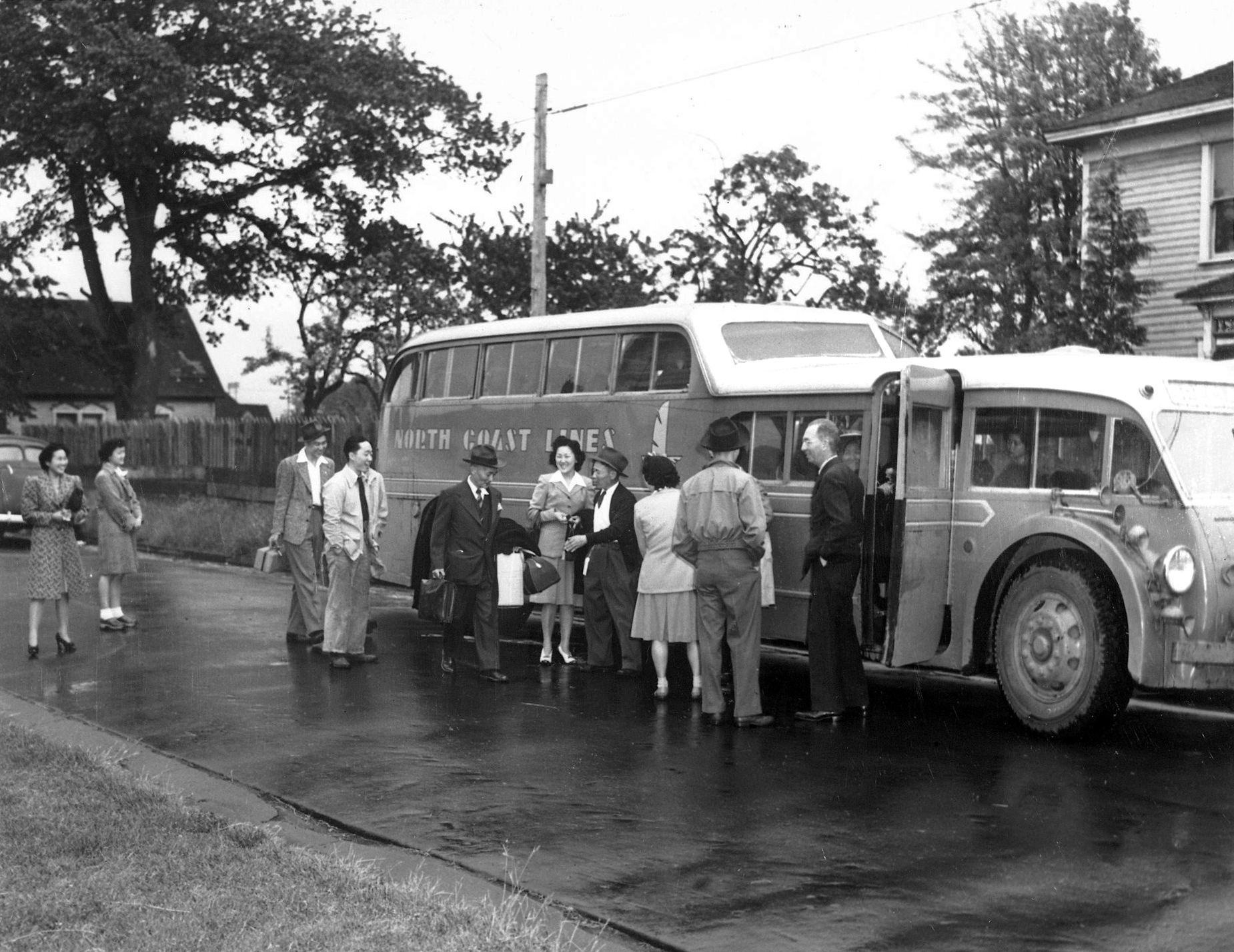 A 1940s photo of several smiling people getting on a bus that&#039;s parked on a tree-lined residential street. 