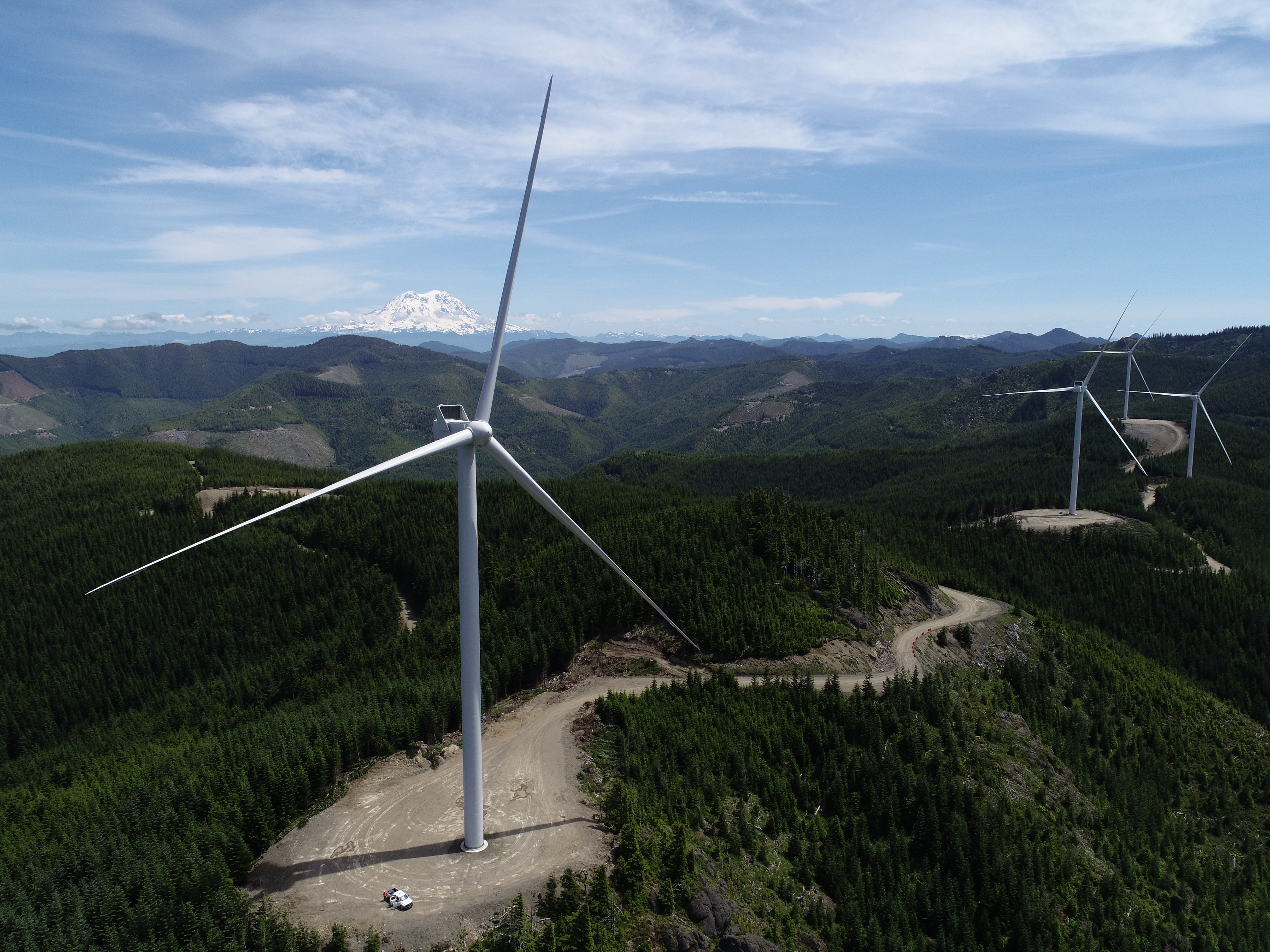Four giant wind turbines sit on a forested mountaintop. For perspective, a truck near the base of one of the three-bladed turbines looks like a tiny toy. 