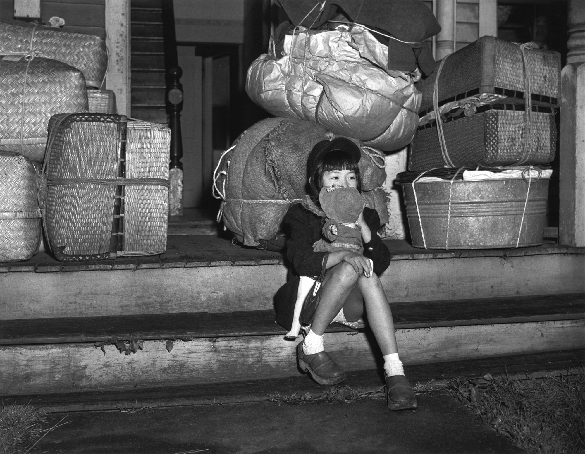 A little girl sits on the front steps of a house, clutching a doll, and surrounded by stuffed wicker baskets, wash tubs and other bundles of belongings ready for transport