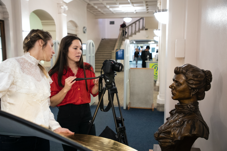 two women stand behind a camera on a tripod, capturing footage of a bronze bust