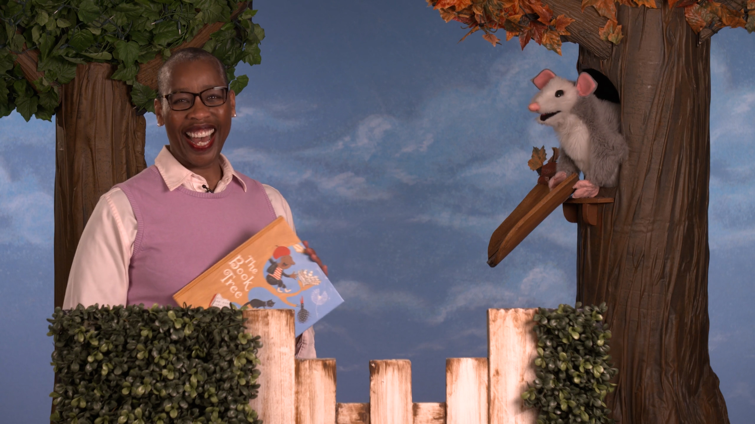A woman on a childrens&#039; television set smiles while holding a book. She stands among make-believe trees and a picket fence. A possum puppet sits in a tree opposite her.