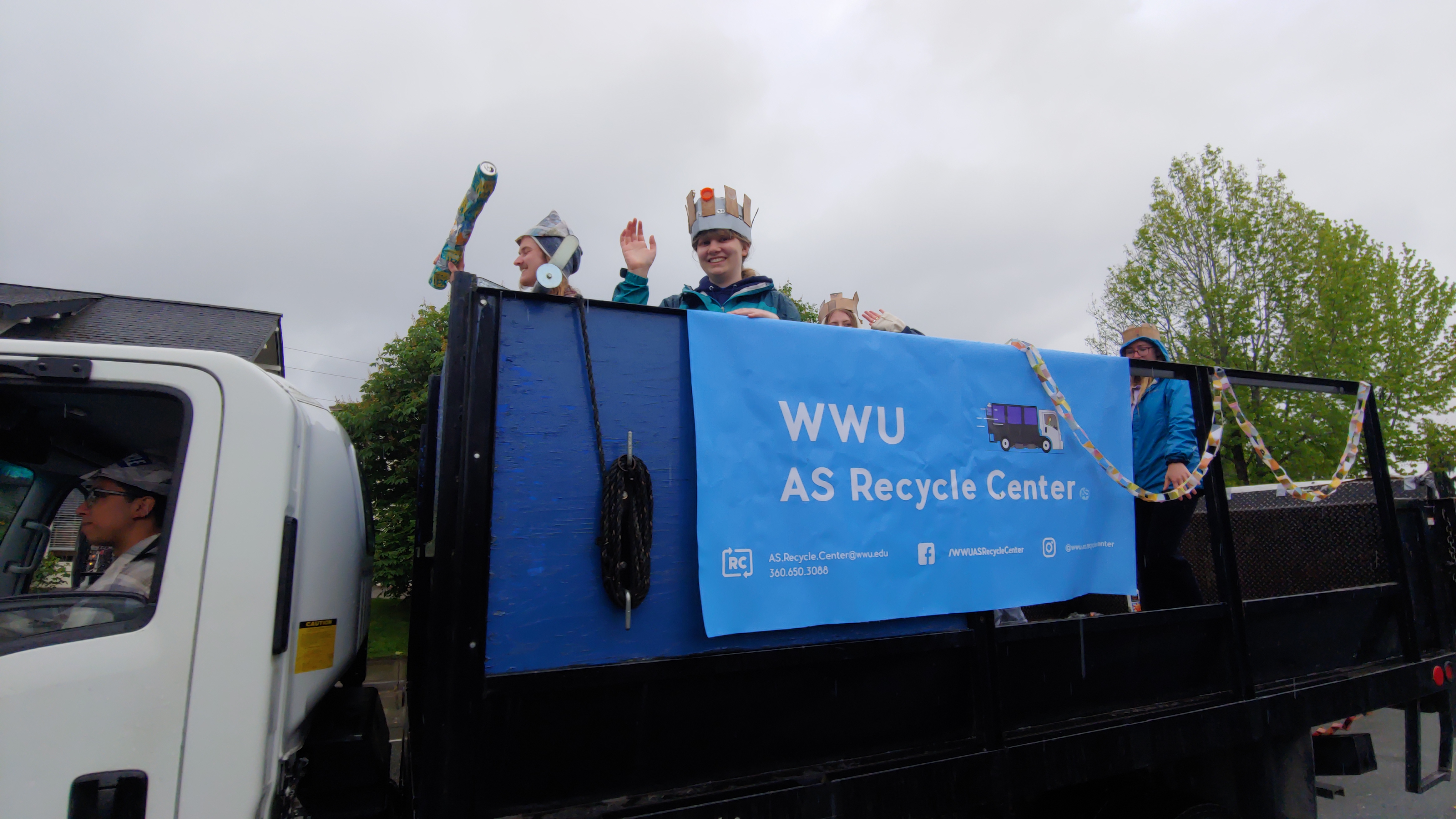 students wave from the back of a truck with a banner, &quot;WWU AS Recycle Center.&quot; The students appear to be wearing hats and crowns made from recycled materials.