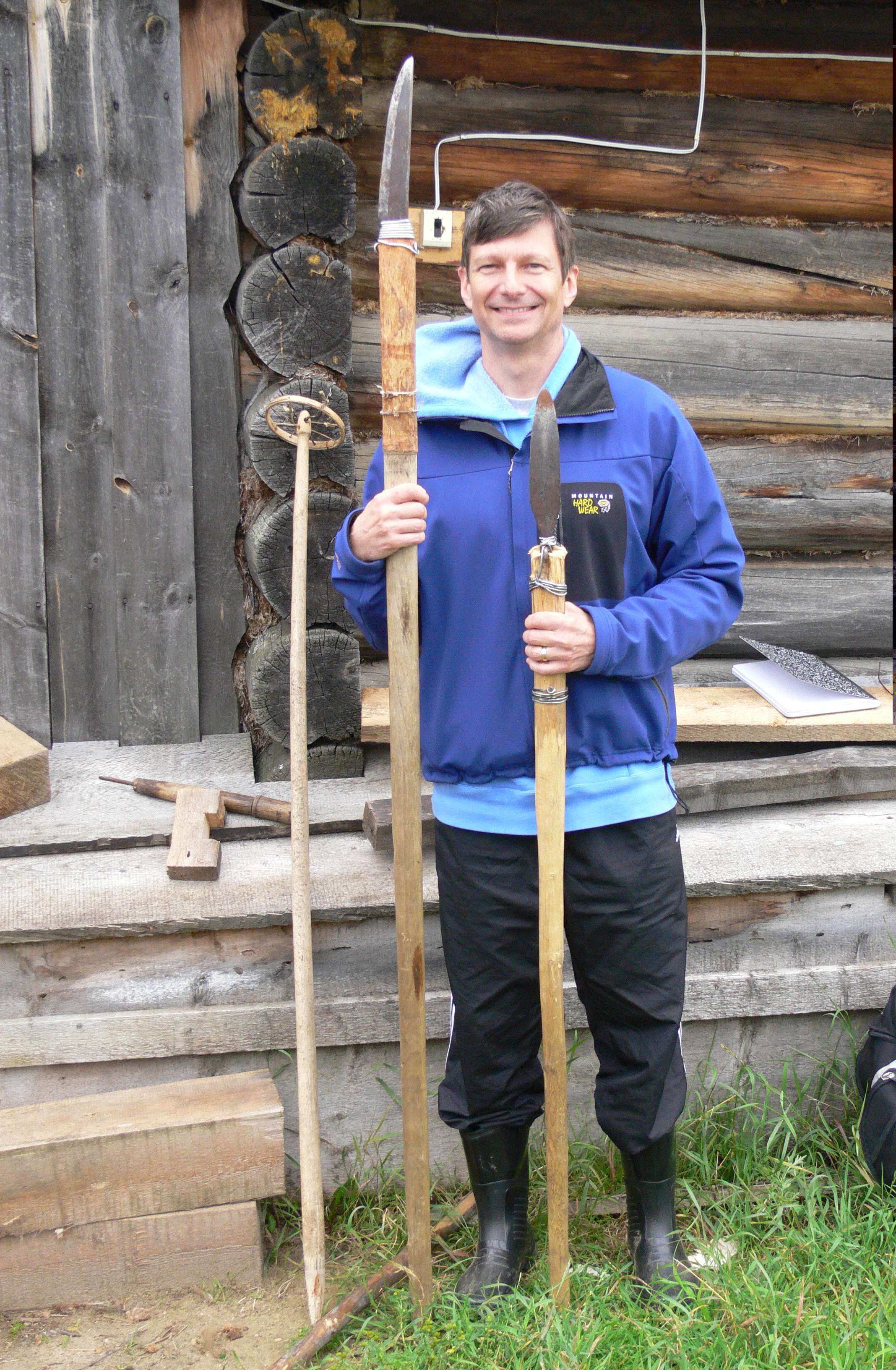 Ed Vajda stands in front of a log cabin and holds two spears