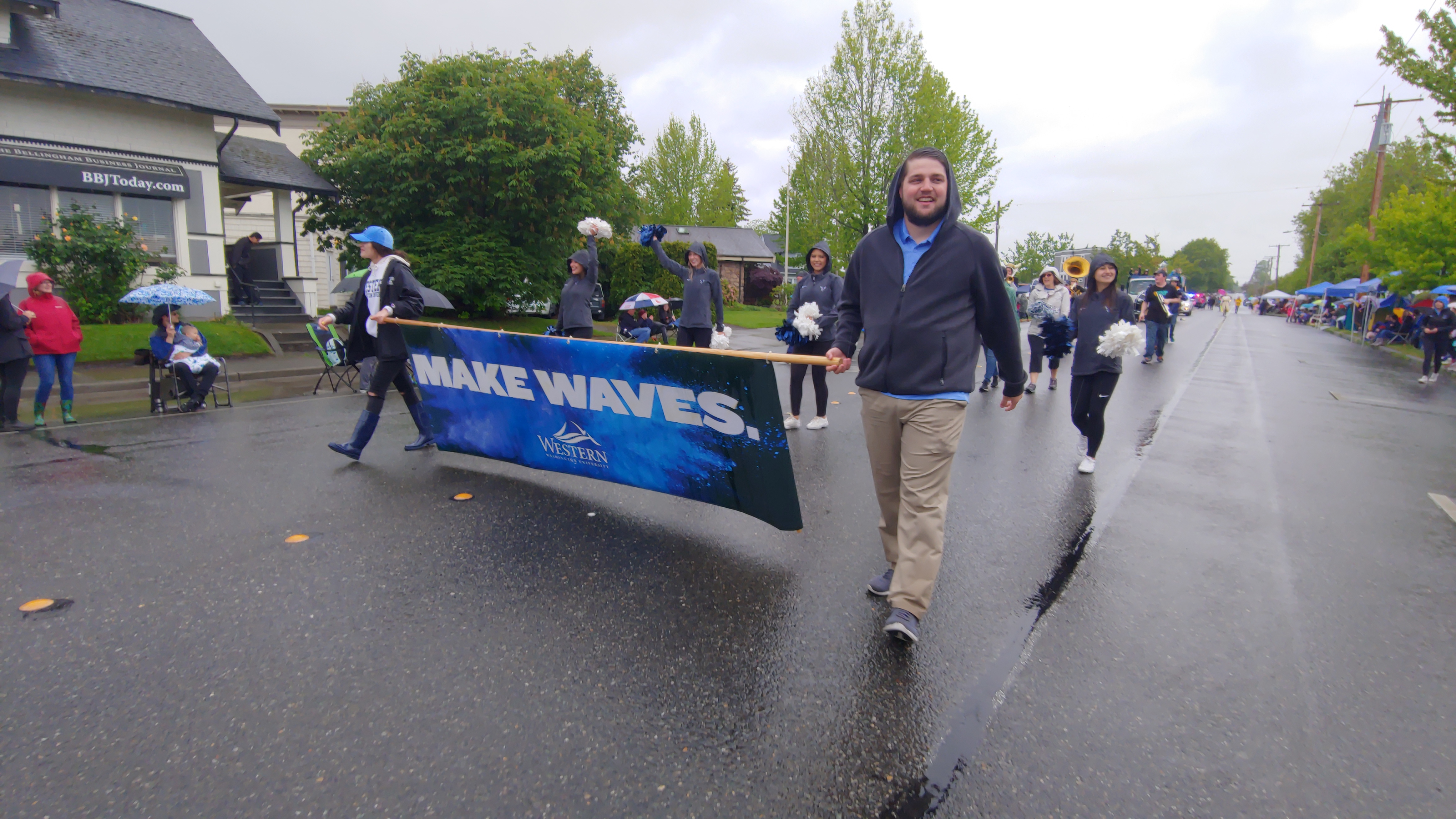 Two people carrying a banner that reads &quot;Make Waves, Western Washington University&quot; lead several people in a parade down the middle of Cornwall Avenue as people holding umbrellas watch from the sides