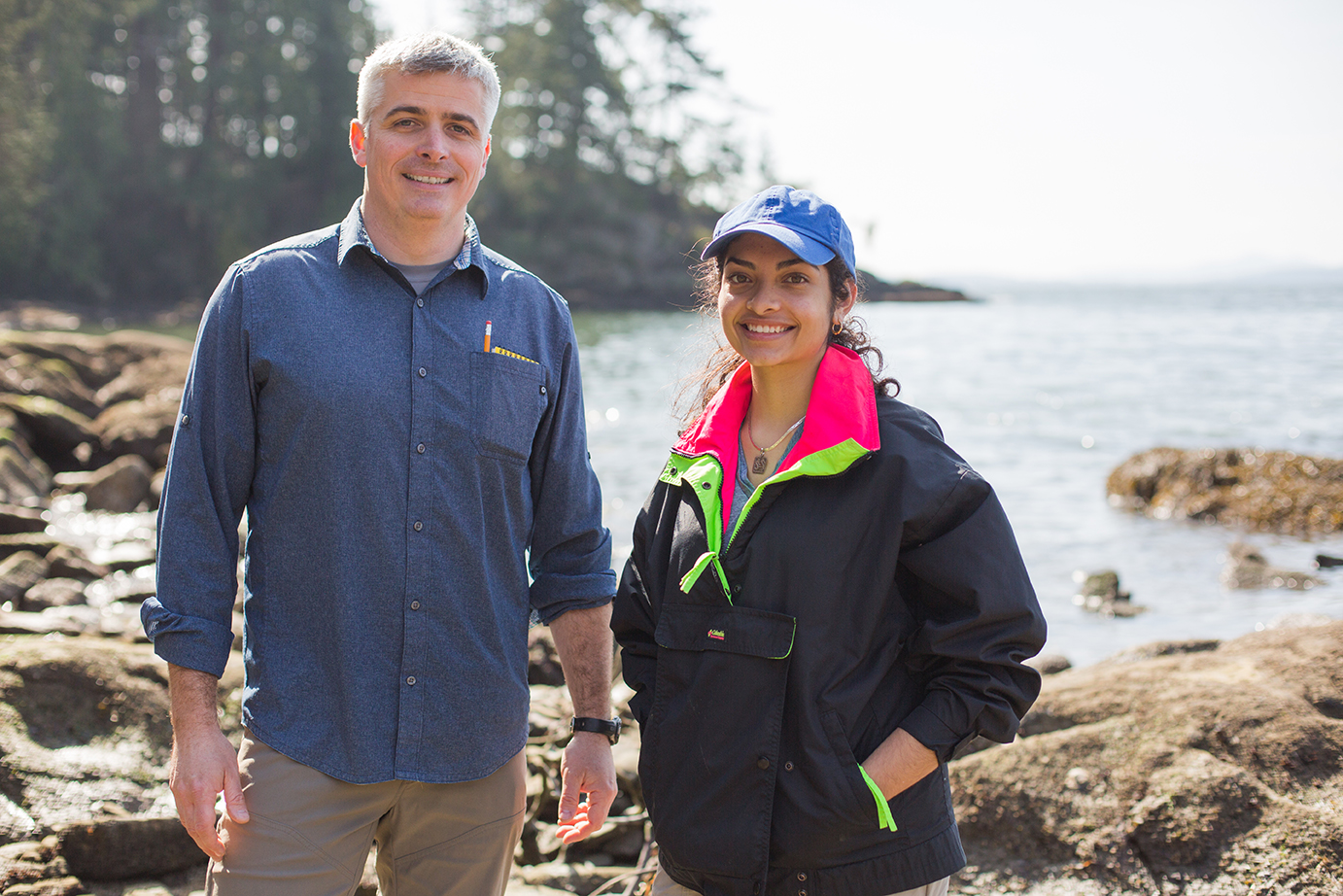 A female student and male professor stand on a rocky beach, smiling at the camera. 
