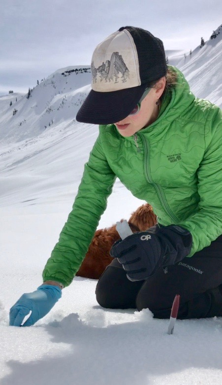 Robin, wearing a green puffy coat and baseball cap, kneels in the snow