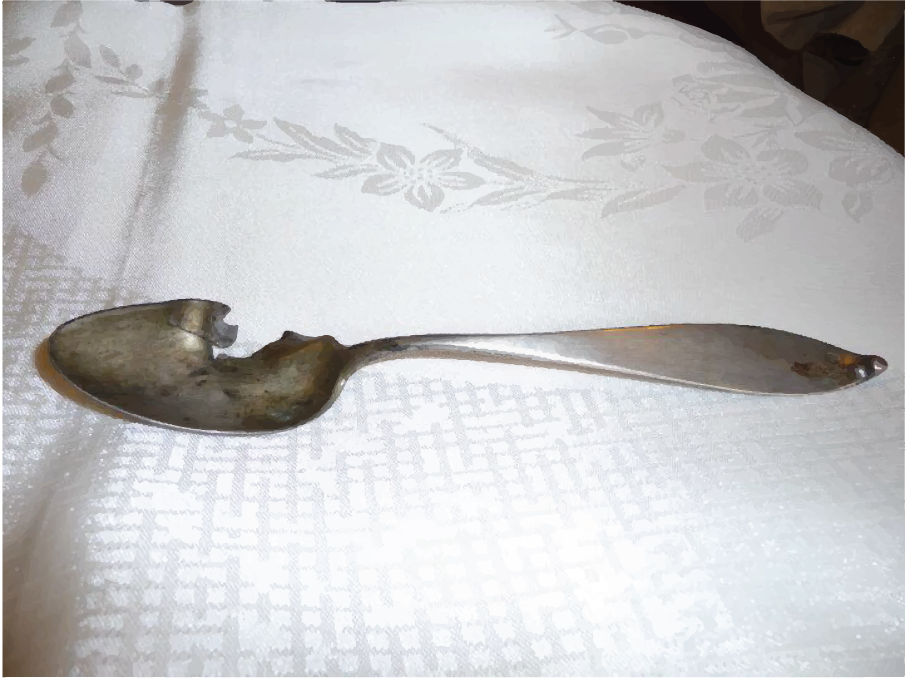 An old, damaged silver spoon sits on a damask table cloth
