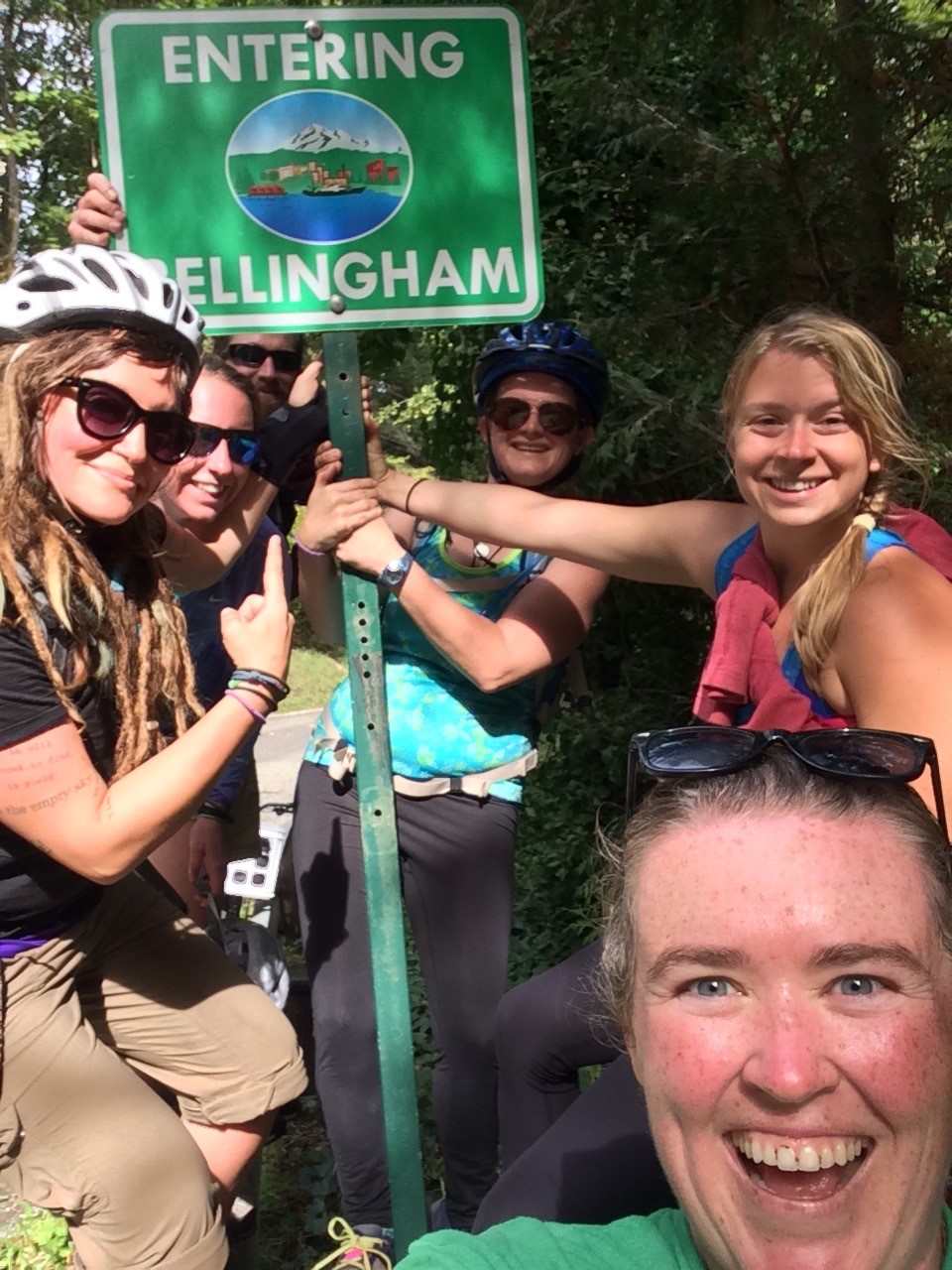 Six smiling people surround a sign that reads &quot;Entering Bellingham.&quot;