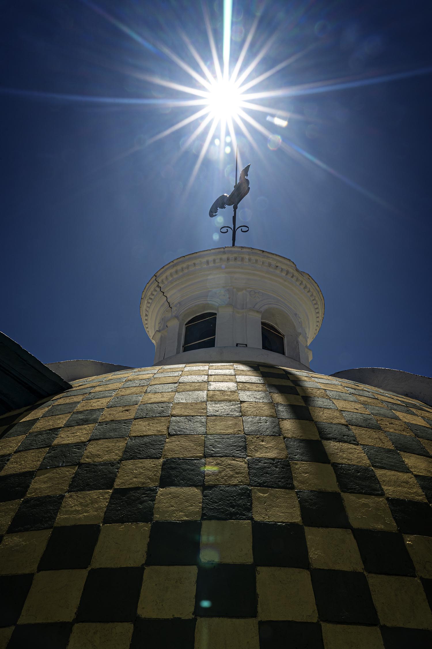 Atop a domed building with a black and white checkered roof sits a rooster weather vane, haloed by the sun