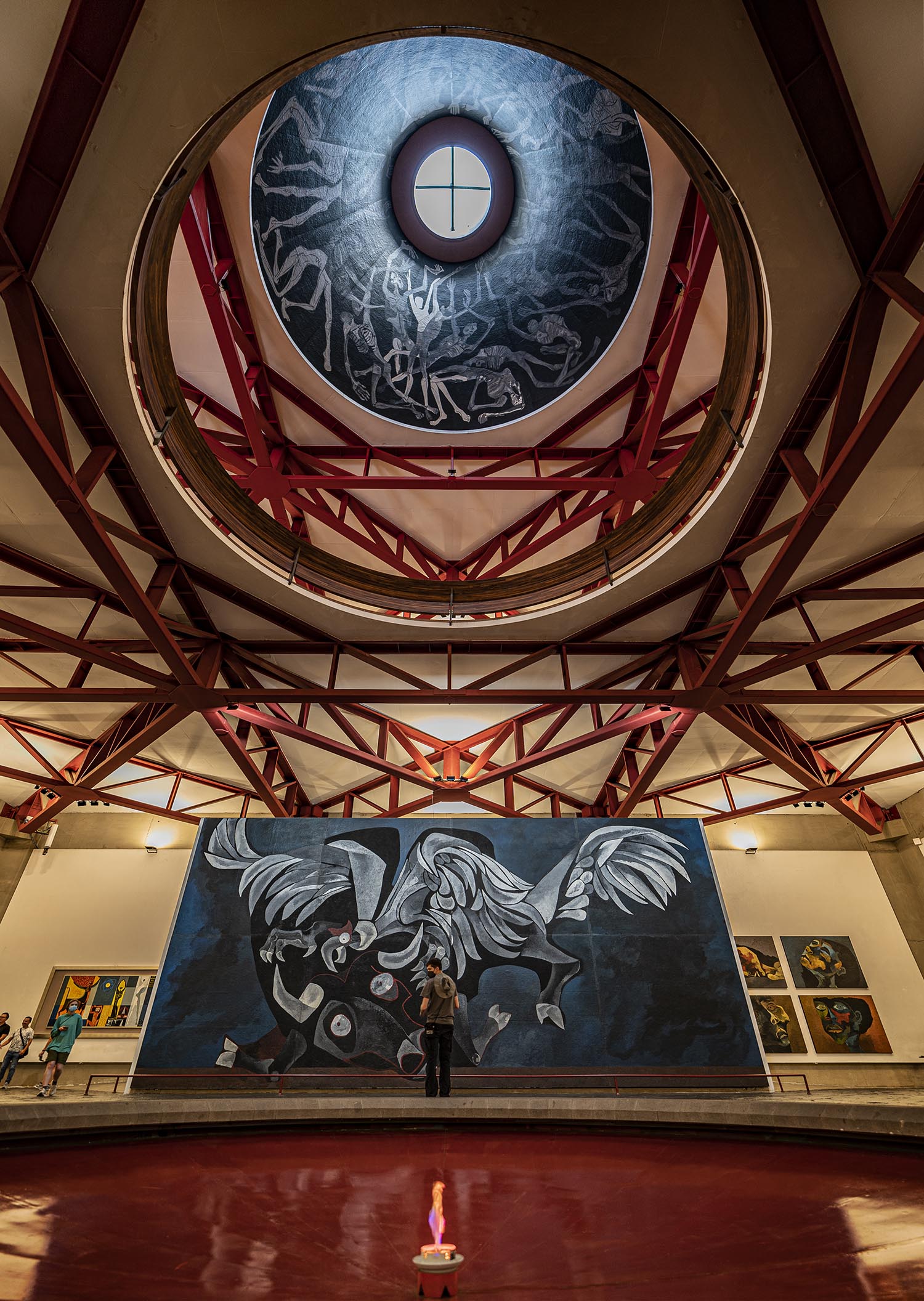 A student standing in front of a large, black and white abstract paining of a winged bull. Overhead red scaffolding surrounds a domed mural of skeletal figures in various states of contortion.