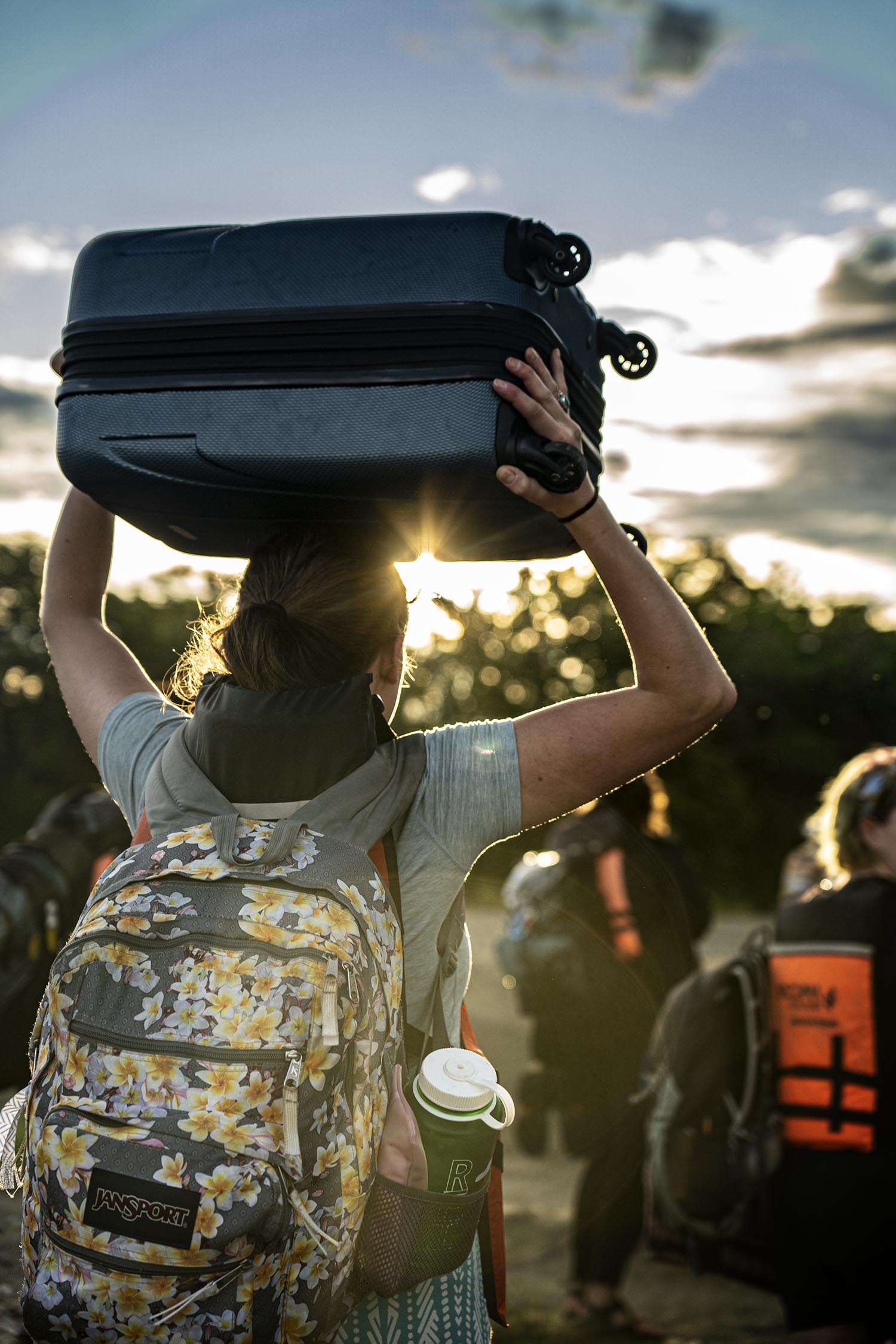 A student carrying a suitcase on their head, silhouetted by the setting sun