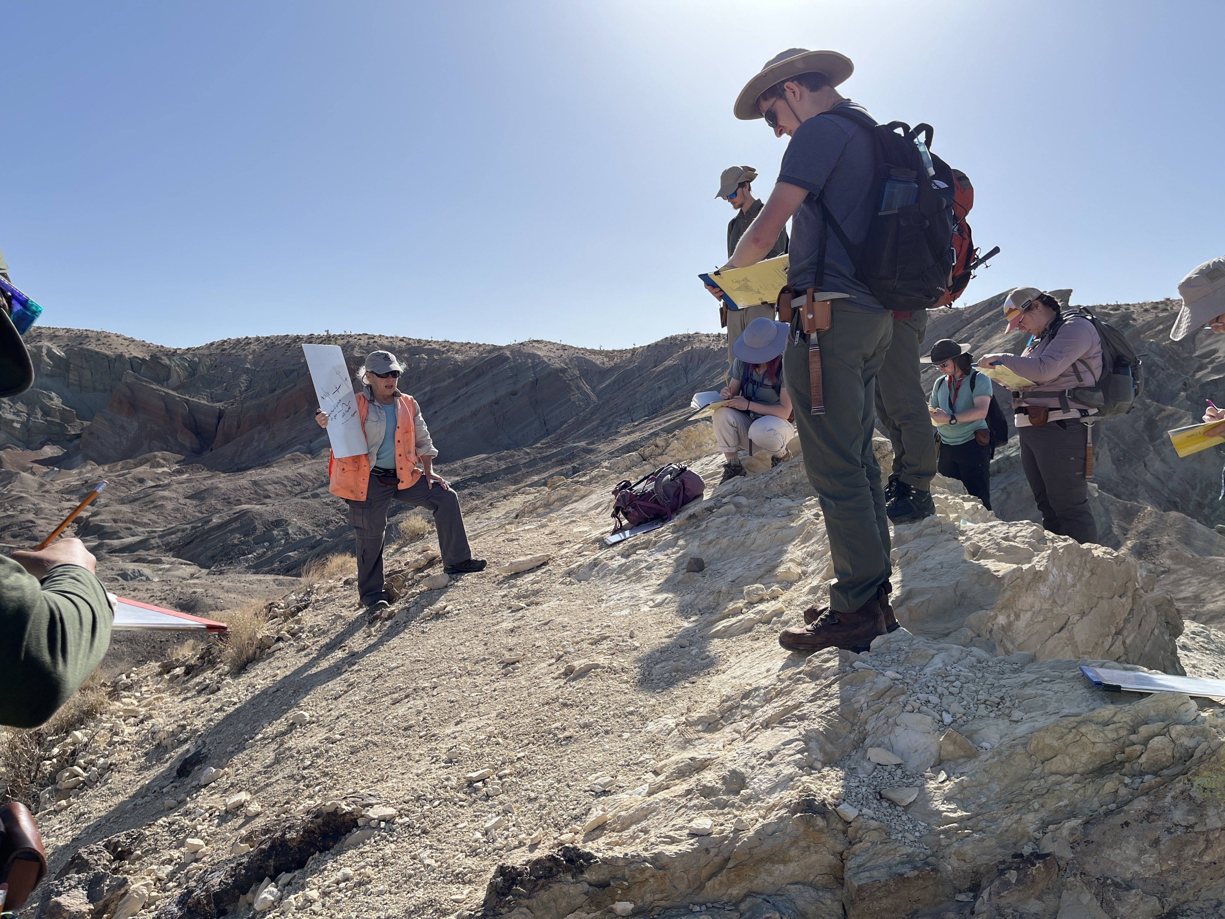 Geology Professor Liz Schermer holds a white board as she lectures on a rocky slope, surrounded by students 