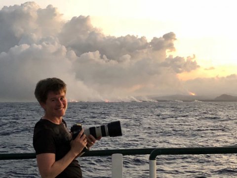 Jackie Caplan Auerbach holds a camera on the deck of a ship, volcanic steam and flames rising from the sea behind her