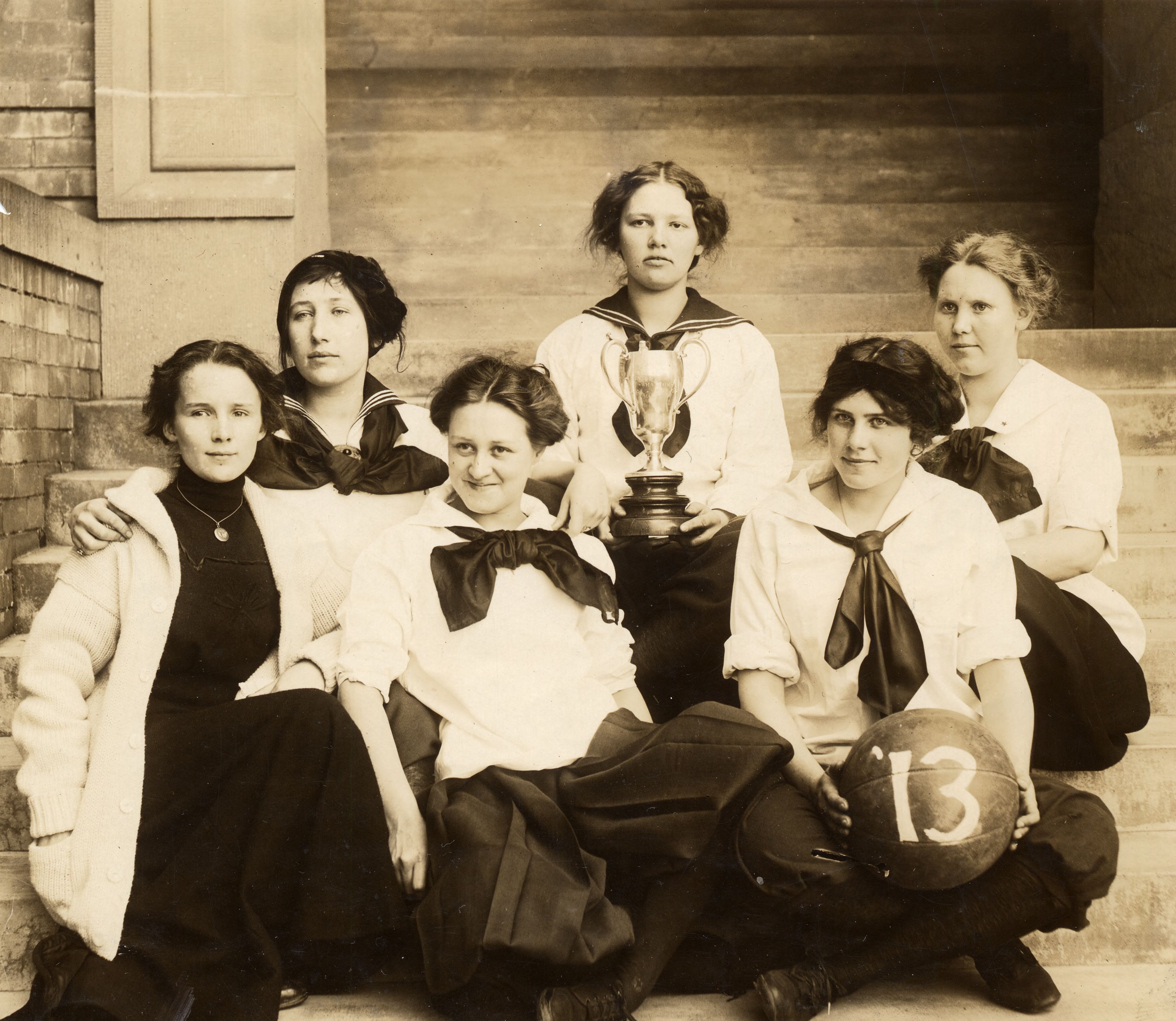 Six students sit on the steps of Old Main in 1913, displaying a trophy cup and a basketball.