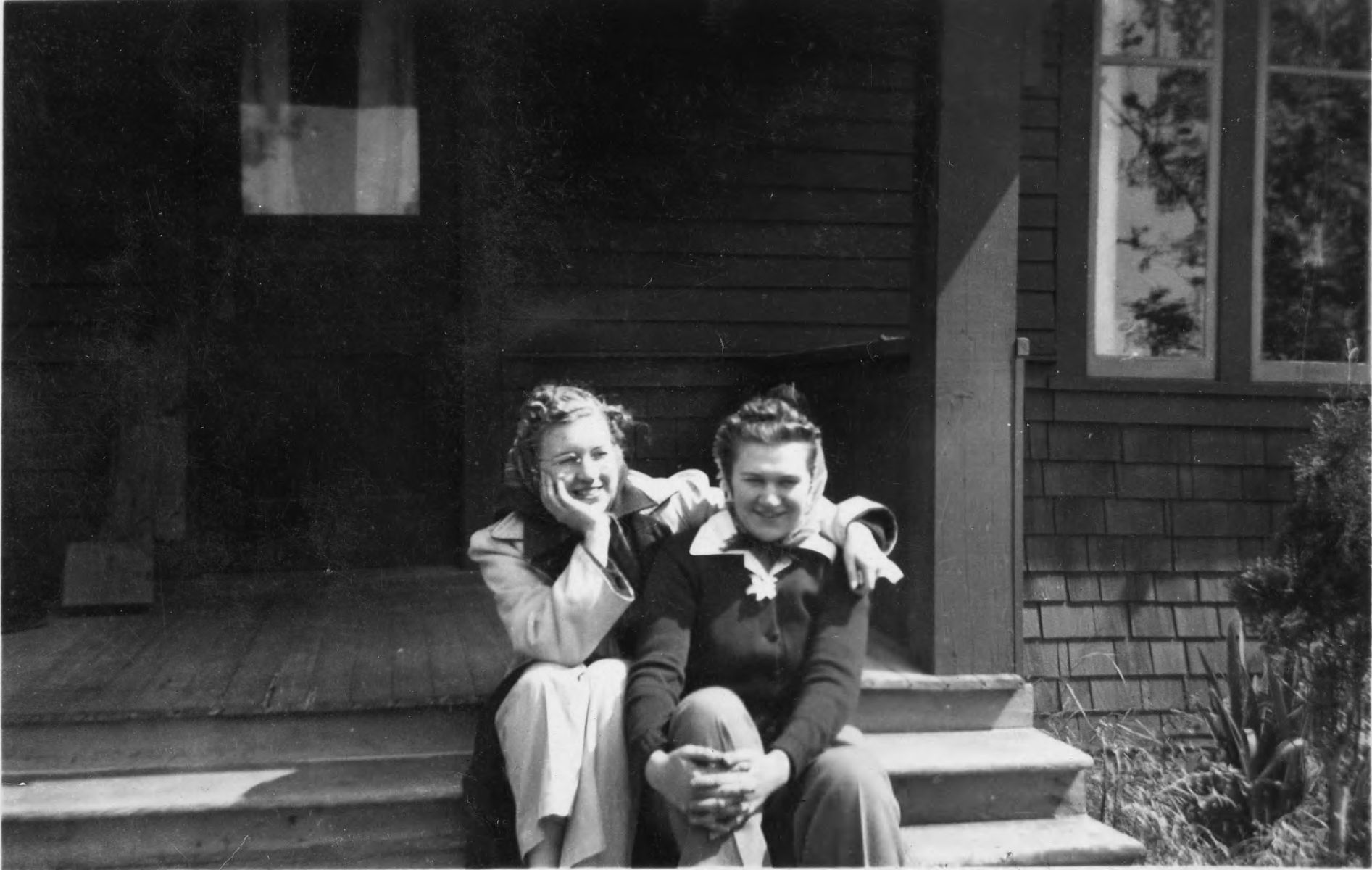 Two women sit next to each other smiling on the porch steps of Viqueen Lodge in 1940