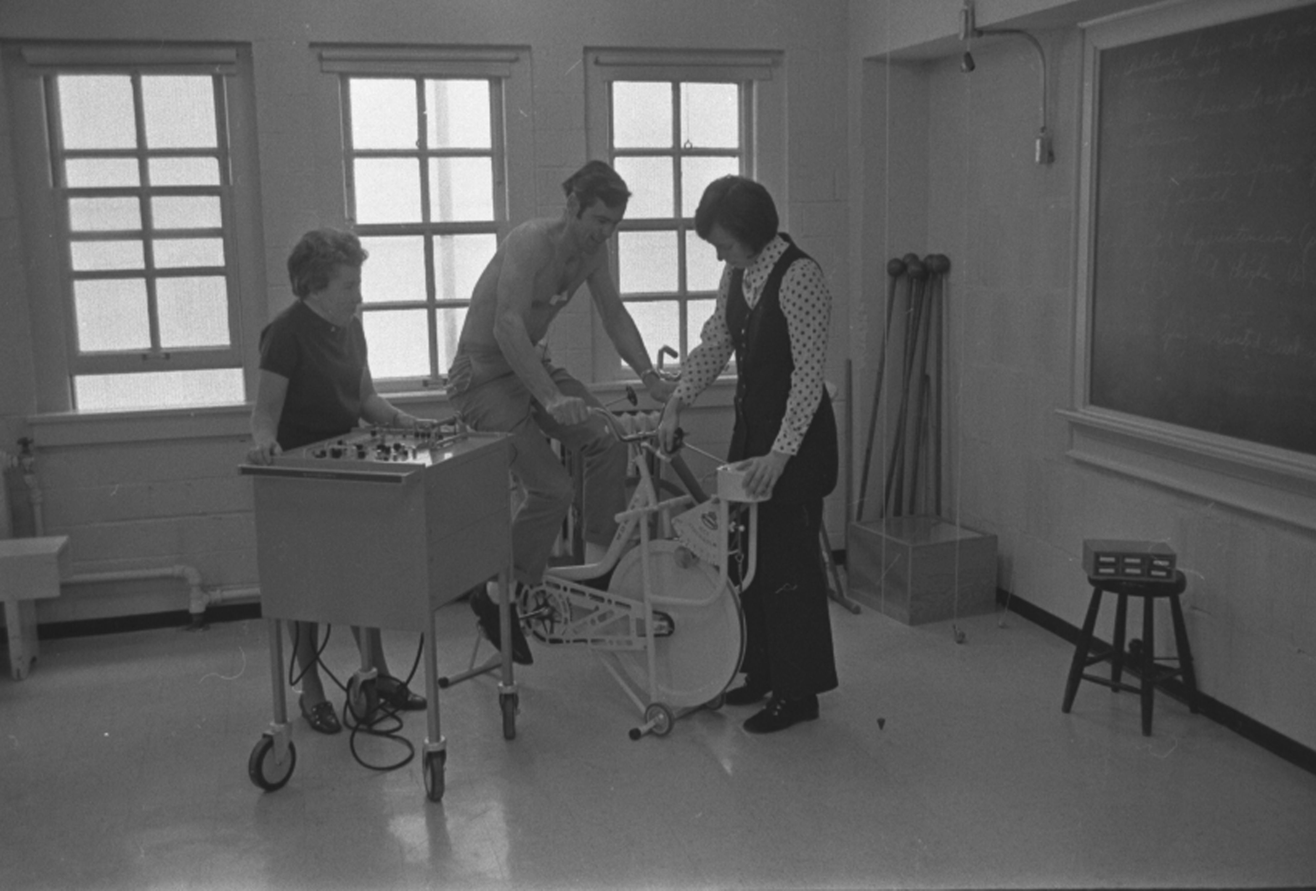 Chappelle Arnett and another person watch a student on a stationary bicycle that is attached to some sort of recording machine in 1971.