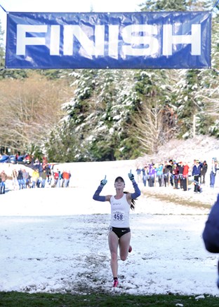 Sarah Porter passes the finish line on a snow-covered trail, smiling and pointing skyward with both hands. 