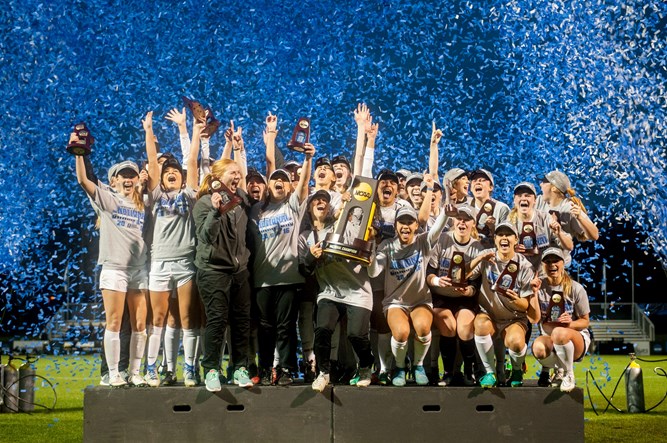 The women's soccer team stands on a podium celebrating and holding championship trophies as blue confetti rains down. 