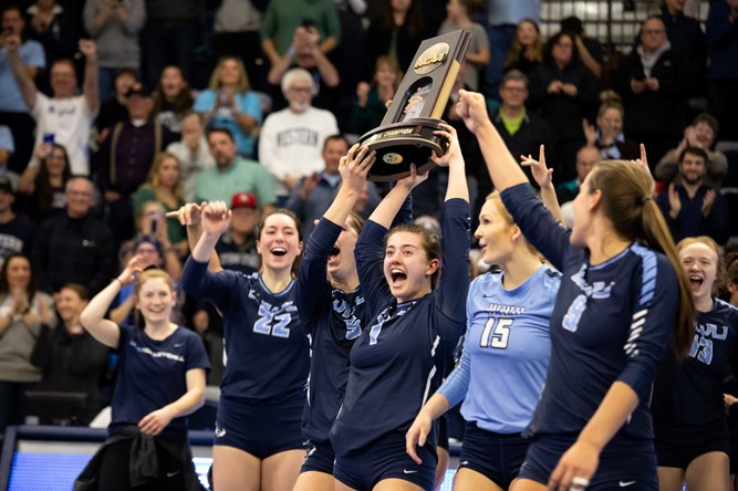 Women's volleyball team members celebrate on the court, hoisting a large NCAA trophy. 