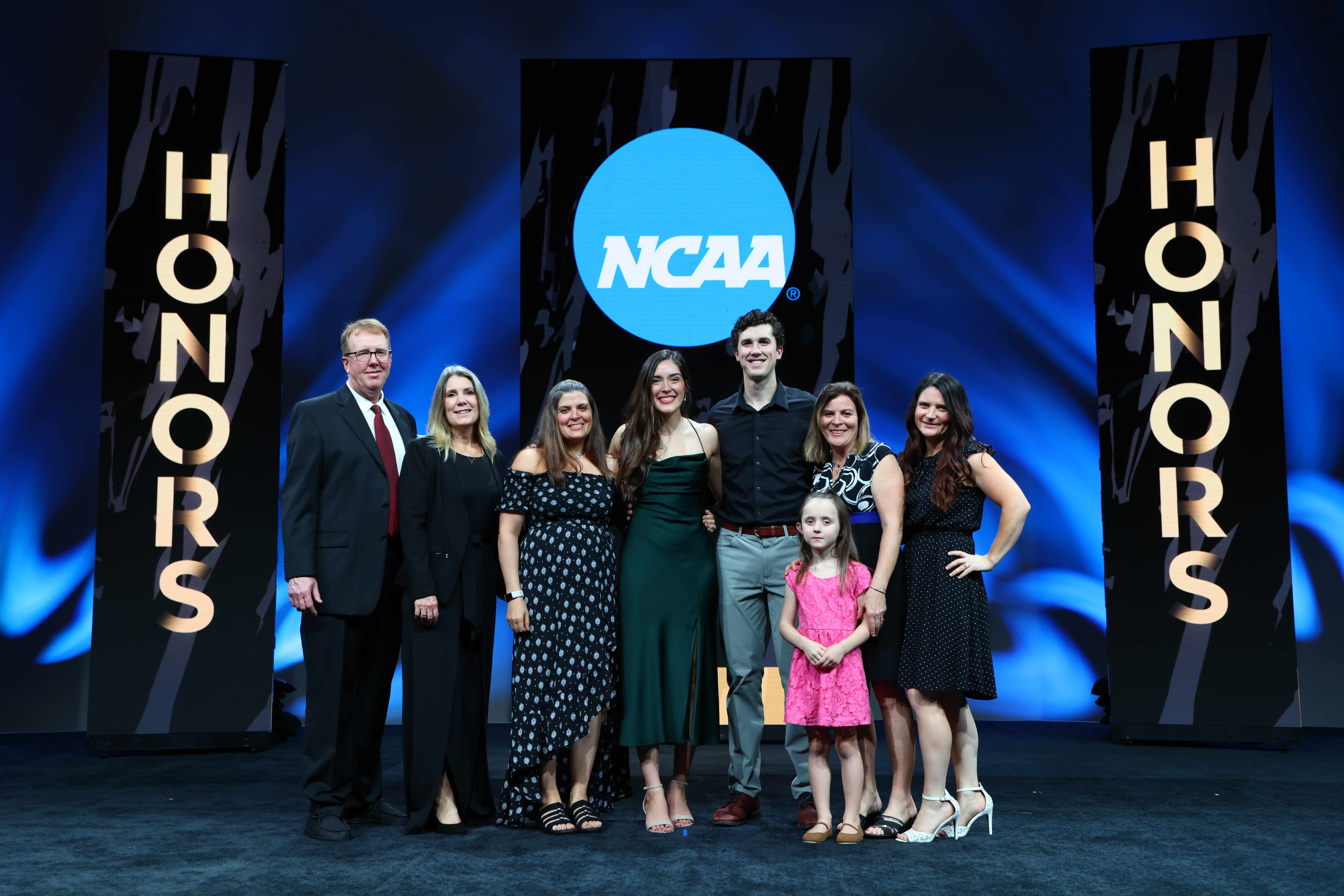 Gracie, wearing a fancy green dress, stands with family members on a stage at the NCAA Honors event. 