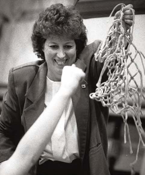 Lynda Goodrich smiles, holding a basketball net cut from a hoop after a win. 
