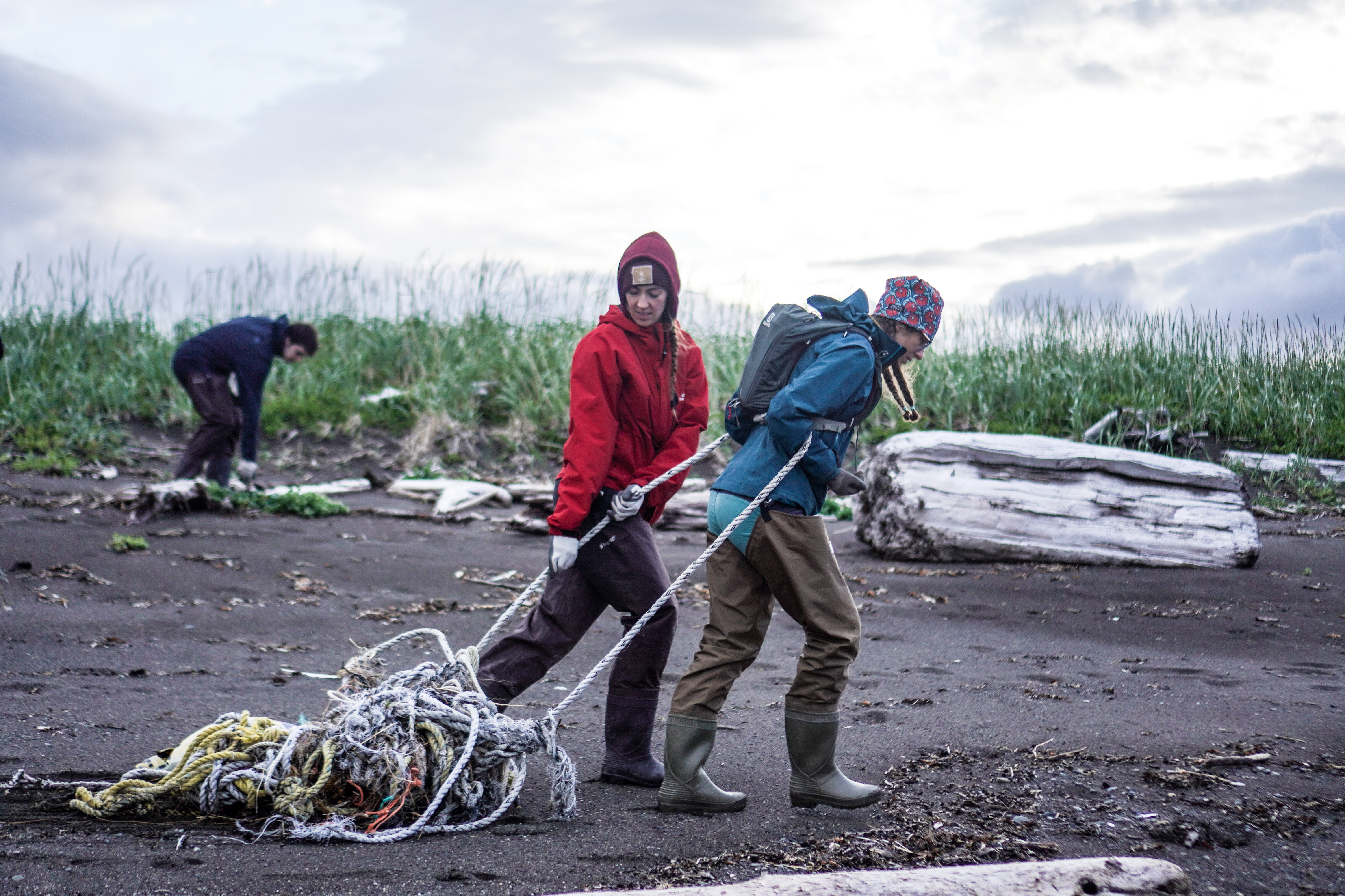 Two people walk across a beach, pulling a tangle of ropes. Another person in the background is picking up plastic trash near some drift wood