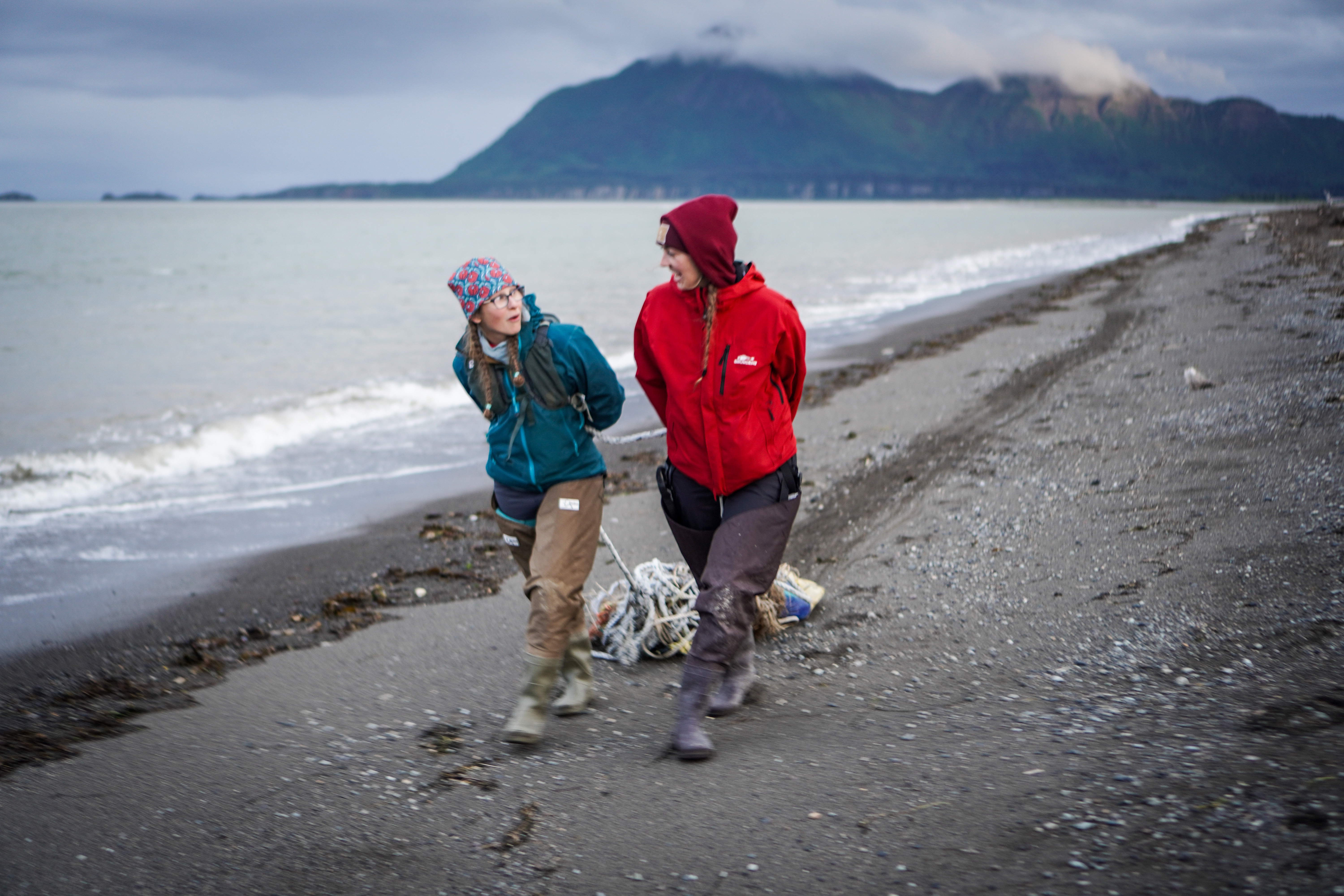 Two people walk down a beach in Alaska, talking to each other as they haul a tangled line of buoys behind them. 