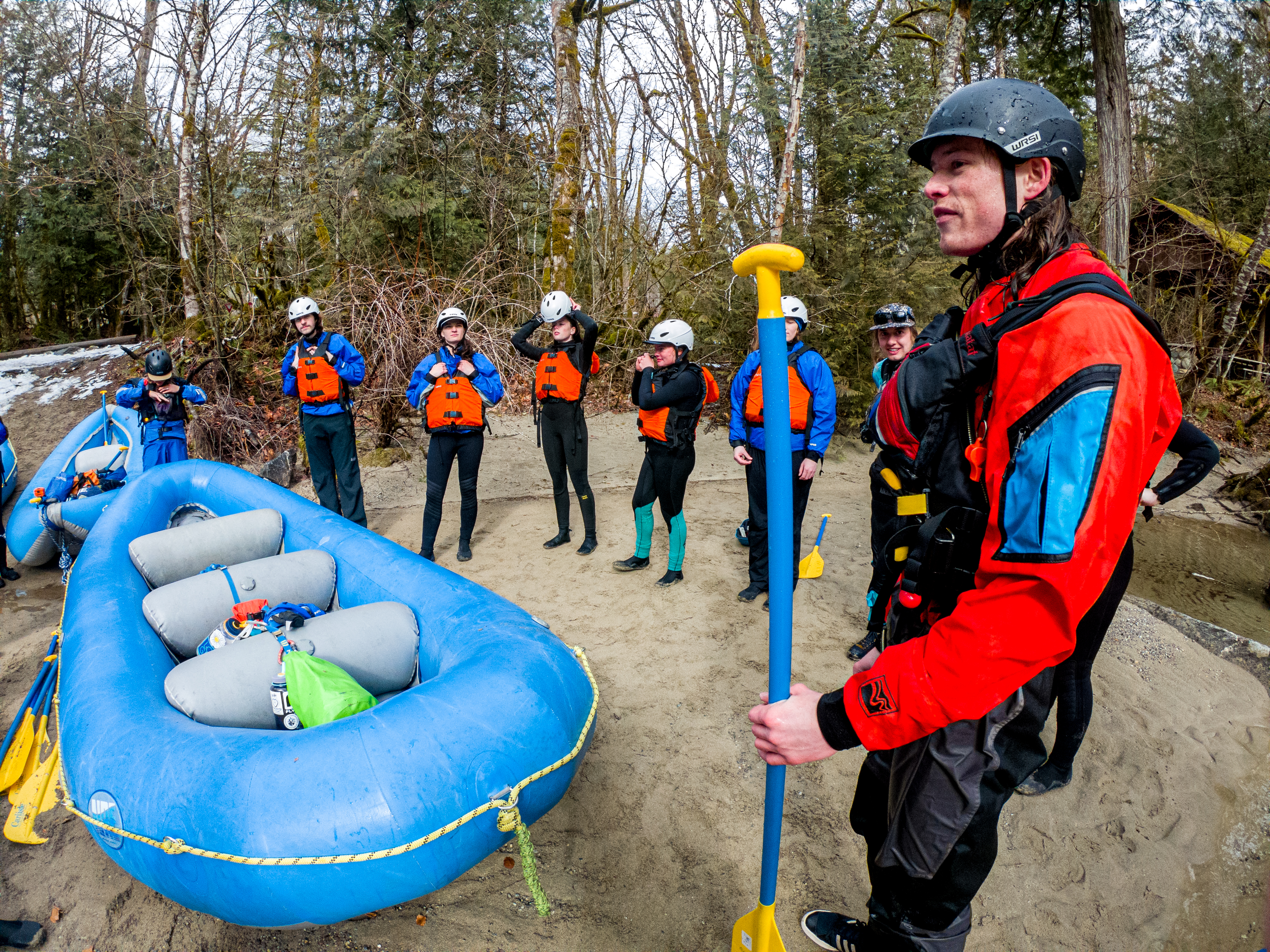 students line a river bank, suited up and ready to put their rafts in the river
