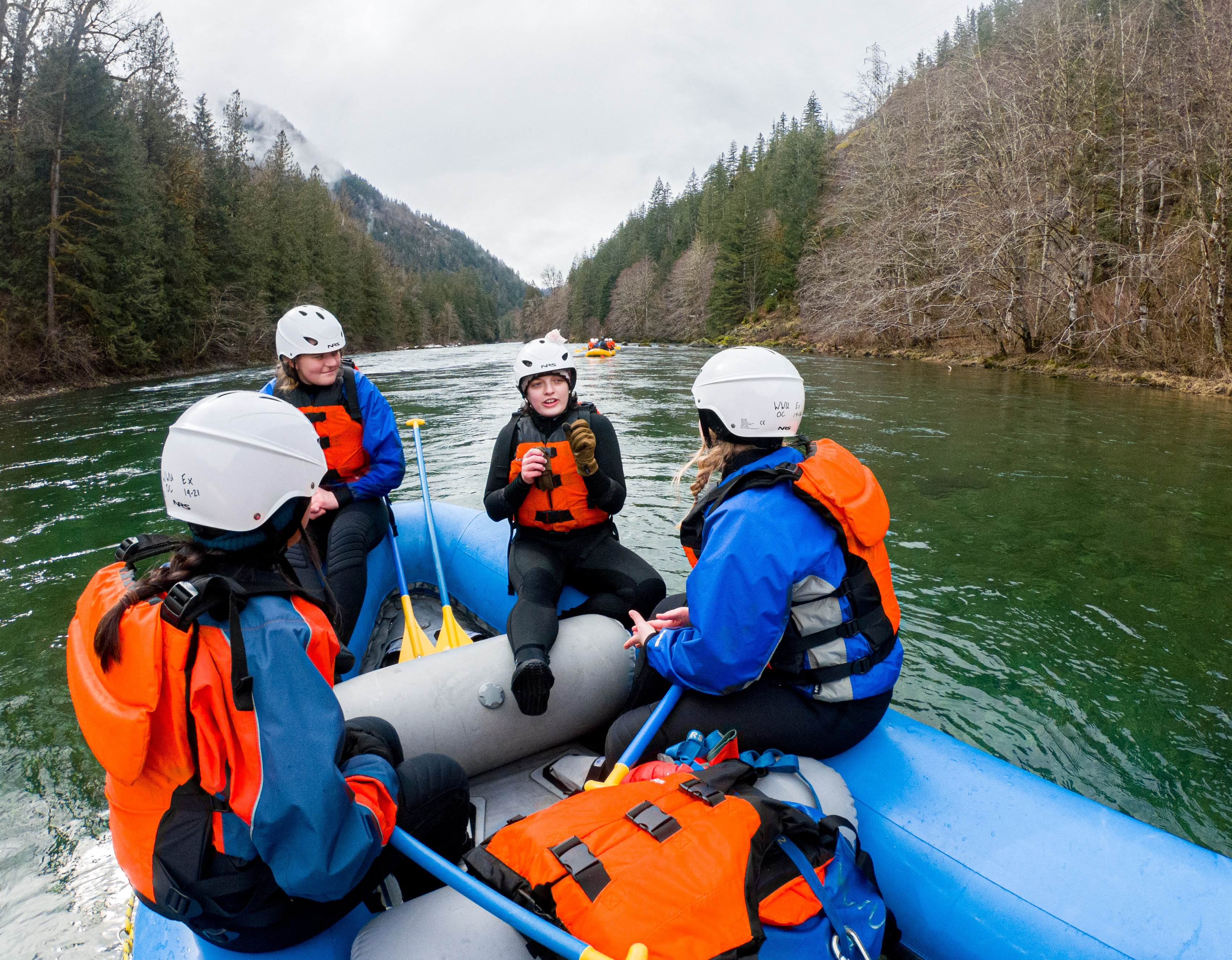 Three students listen to a fourth student while they float in the raft down the river, trees lining the riverbanks. 