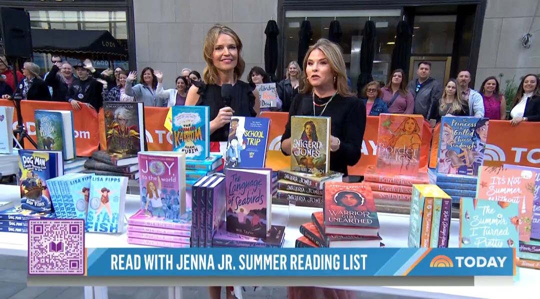 Two Today Show hosts stand on a New York Street behind a table filled with kids' books on display. 