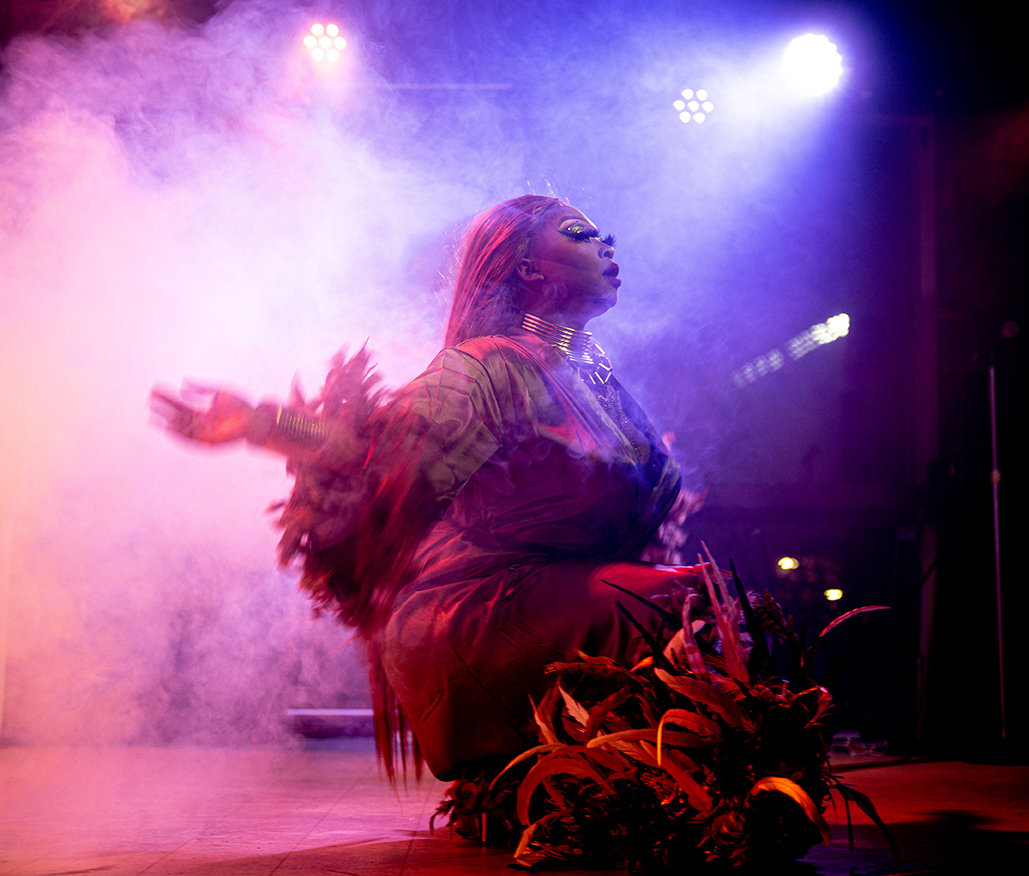 A drag performer sits low on the stage, surrounded by pink fog and blue light