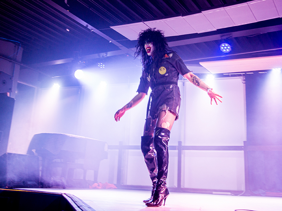 A drag performer with white makeup red lipstick and a black rocker wig, reminiscent of KISS in the 1970s, performs in black patent thigh-highs and a ripped, sequined dress. 