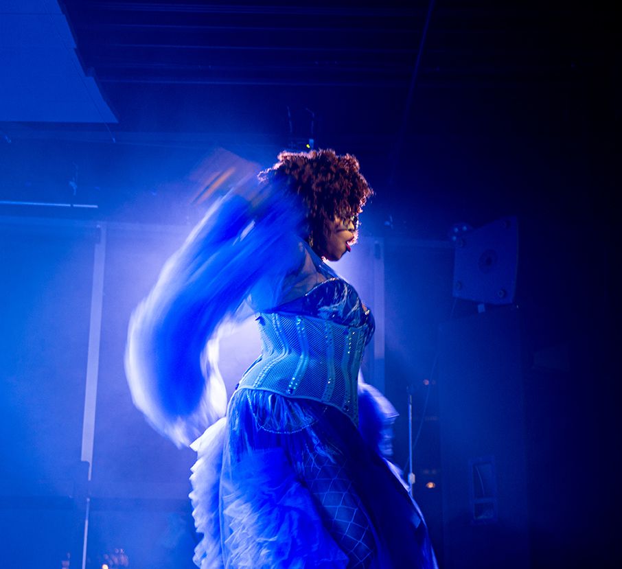 A drag performer in a blue corset dress with fur-trimmed sleeves and a frothy tulle skirt dances on stage, her arms blurred in motion. 