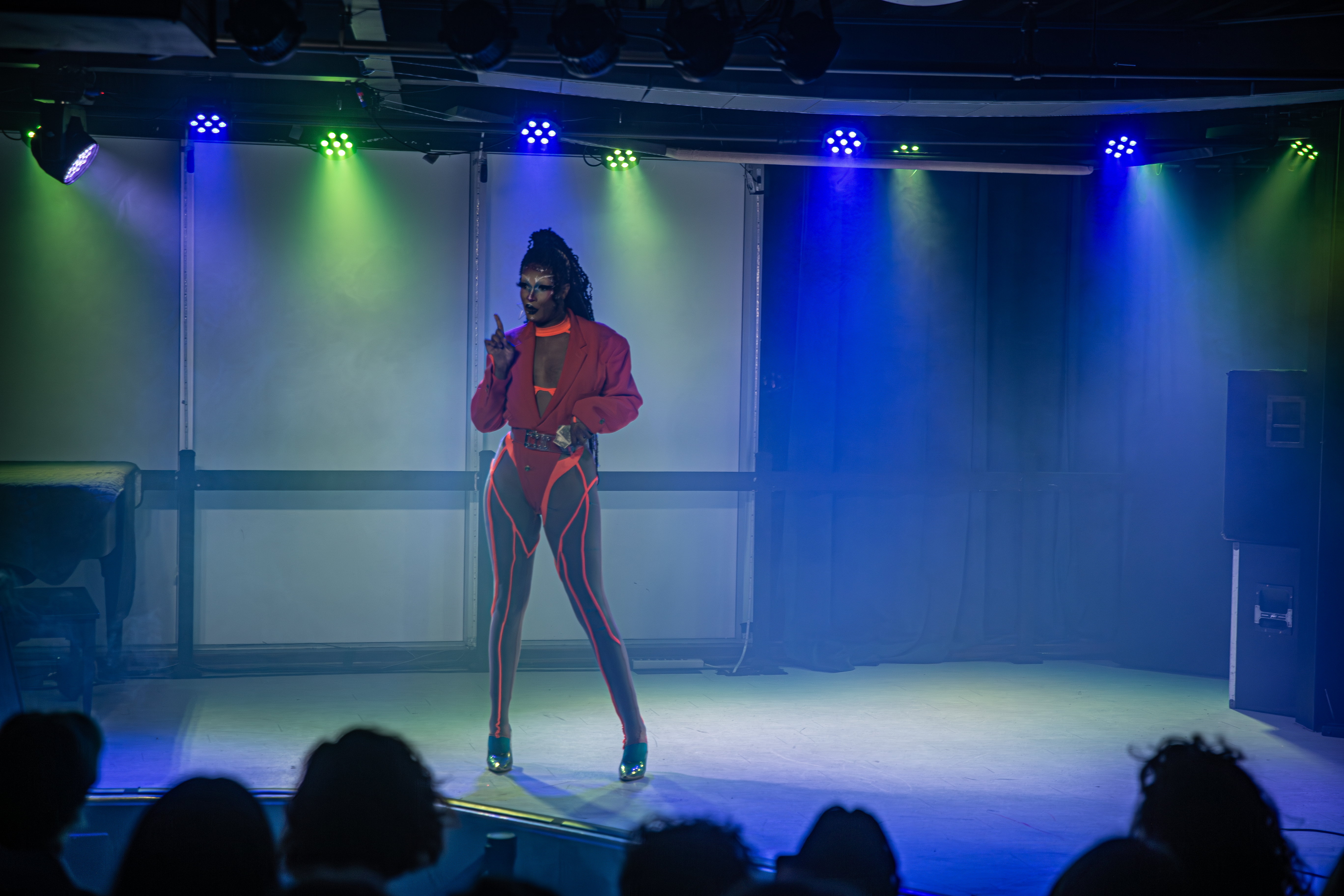 A drag performer wearing neon orange and red speaks to the audience