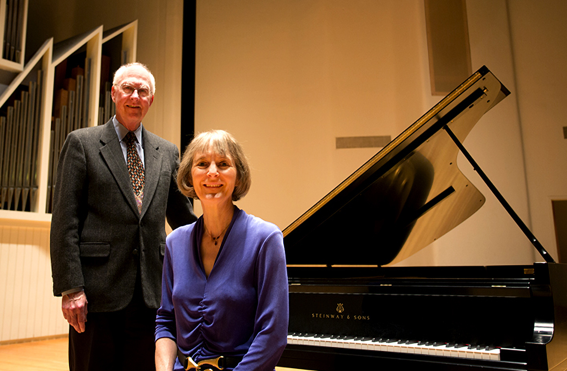 Ford Hill stands and Sybil Sanford is seated at a piano bench on stage in the WWU Concert hall