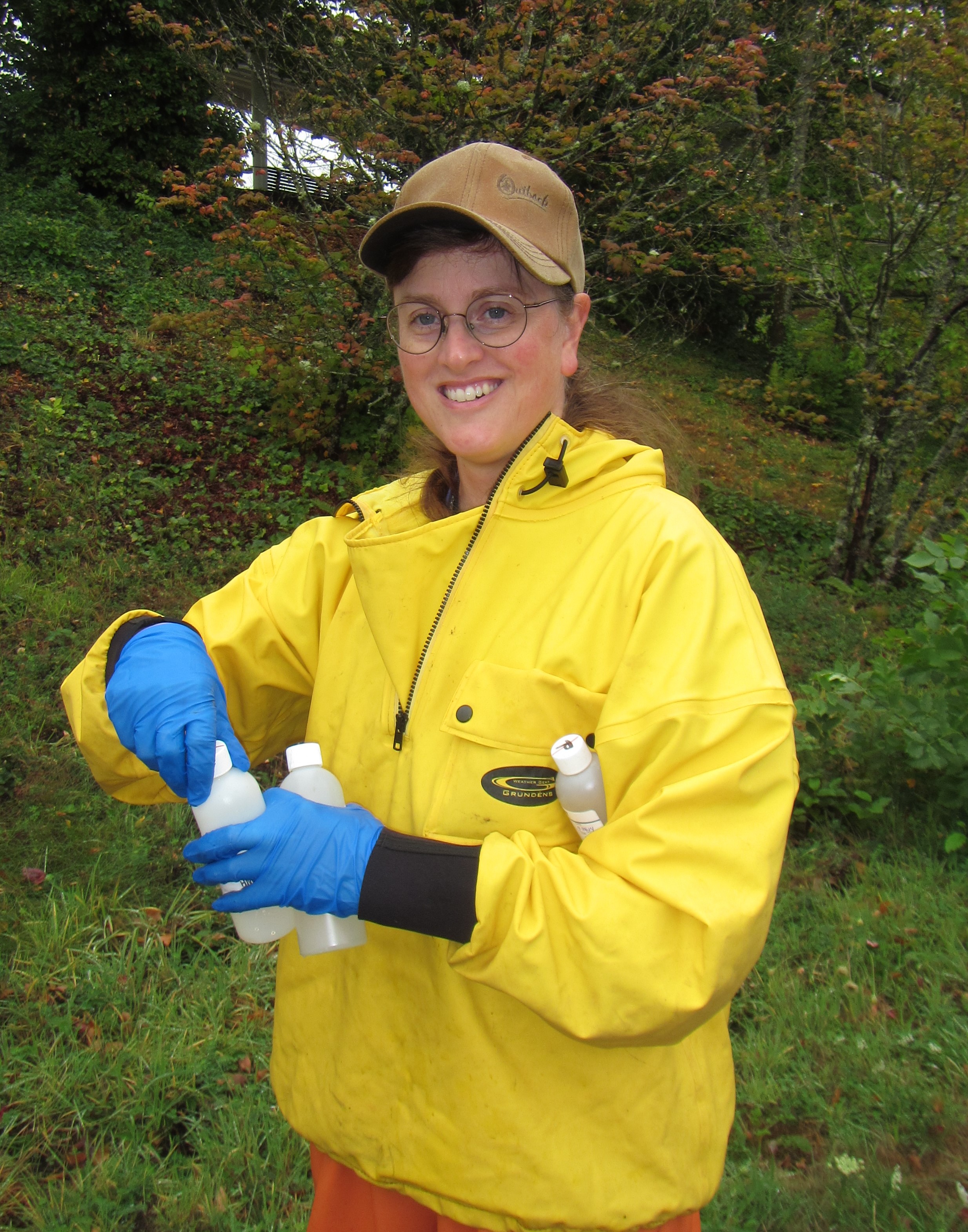 Julann Spromberg, wearing bright yellow waterproof gear, smiles while holding several sample bottles