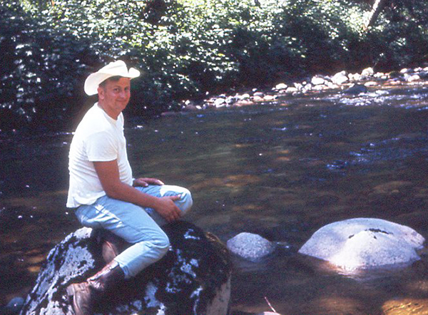 a man smiles, wearing a cowboy hat, white T, jeans and boots, sitting on a rock next to a stream. 