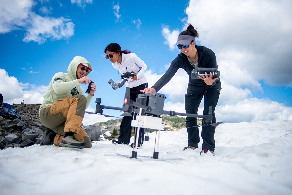 A student crouches down to adjust a piece of equipment on a drone parked on a snow bank on top of a mountain. Two other people are nearby with equipment and watch him work. 