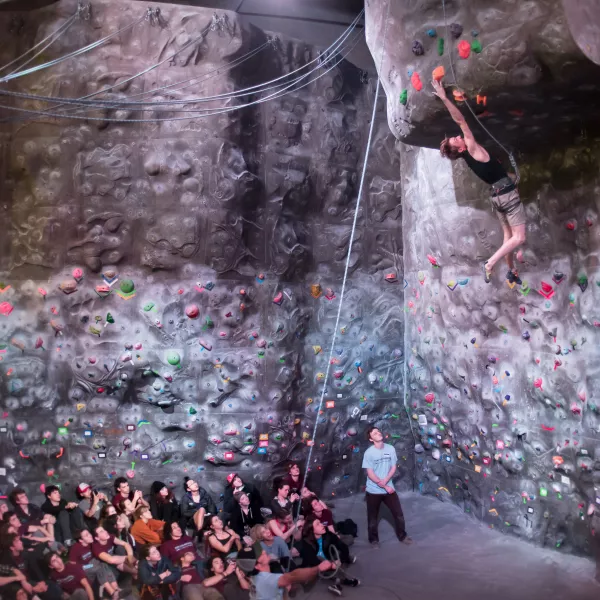 a crowd of students sit on the mat next to a climbing wall, watching one student stretch out his arm toward a handhold above his head