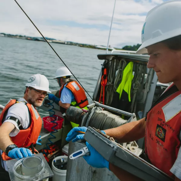 a student holds a clipboard while a faculty member hands him a sample jar on a boat in Bellingham Bay