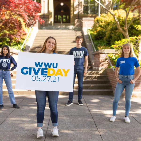Four students wearing WWU t-shirts stand in front of Old Main; one holds a sign with WWU Give Day 05.27.21