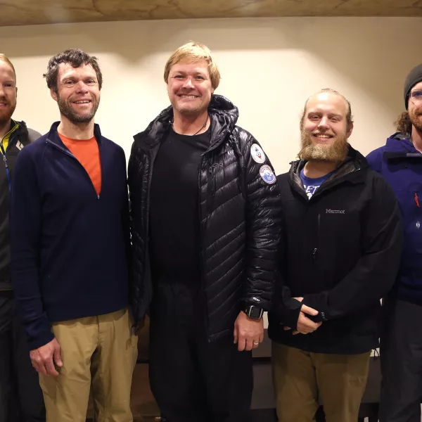 Five men wearing outdoor gear stand inside, smiling and facing the camera