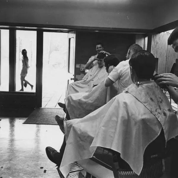 Three students sit in a line of barber chairs while barbers trim their hair. One student sits and waits. 