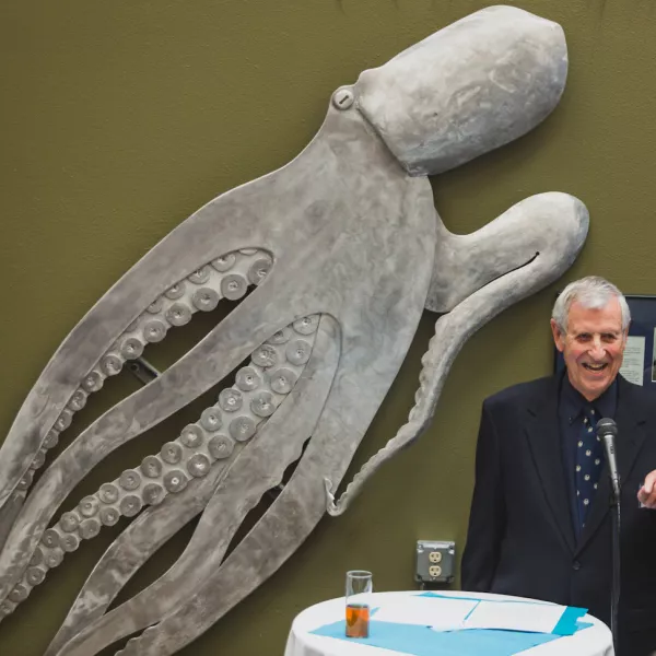 a person speaks at a microphone with a large sculpture of an octopus on the wall behind