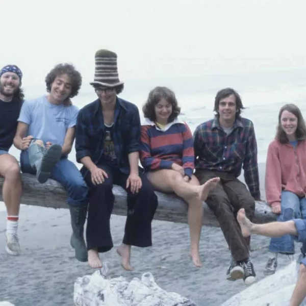A 1970s photo of seven smiling students sitting on a driftwood log on the beach. One of the students is wearing a striped crocheted top hat. 